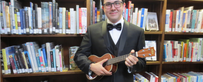 young man in a tux holding a ukulele in front of bookshelves