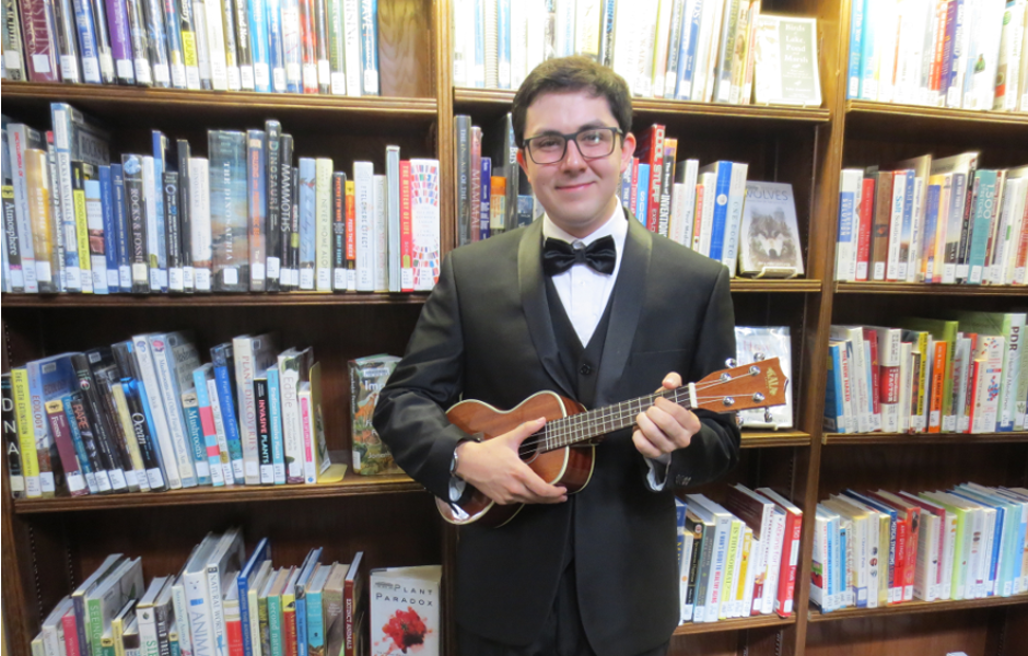 image: young man in a tux holding a ukulele in front of bookshelves